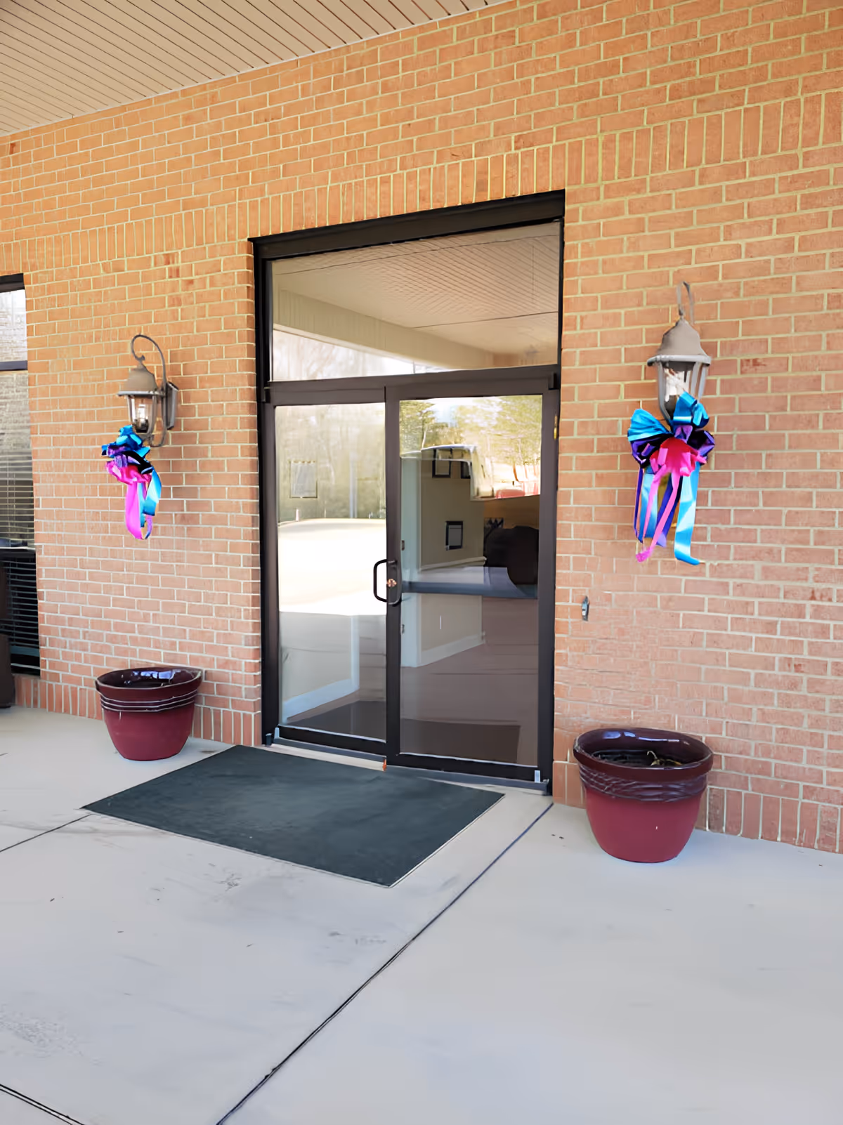 Glass double-door entrance in a brick wall with two large planters and colorful ribbon decorations on wall lanterns.