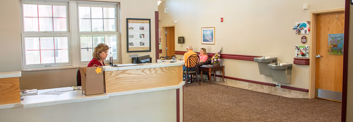 Reception area in Clepper Manor with a woman sitting behind a wooden front desk. Two people are seated on chairs along the wall near a table with a flower arrangement. There are two water fountains mounted on the wall and a door with a welcome sign. Large windows allow natural light into the space.