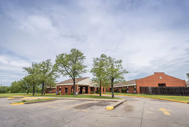 Exterior view of Webb City Health and Rehabilitation Center showing a single-story brick building with a covered entrance, several trees, and an empty parking lot under a cloudy sky.