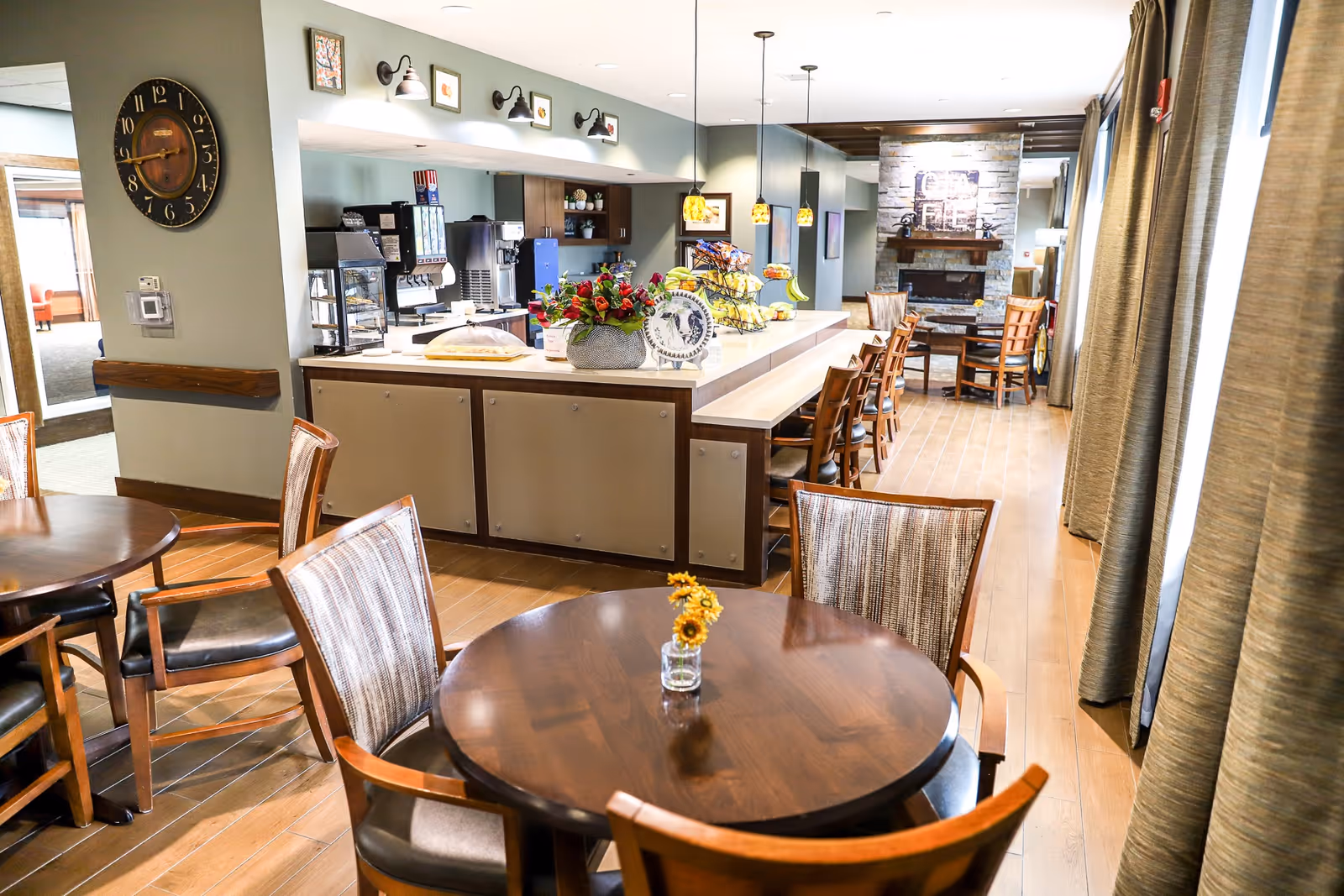 A bright dining area in a senior living facility featuring round wooden tables with chairs, a long counter with bar stools, pendant lights hanging above, and a stone fireplace in the background. The room has large windows with curtains allowing natural light to fill the space.