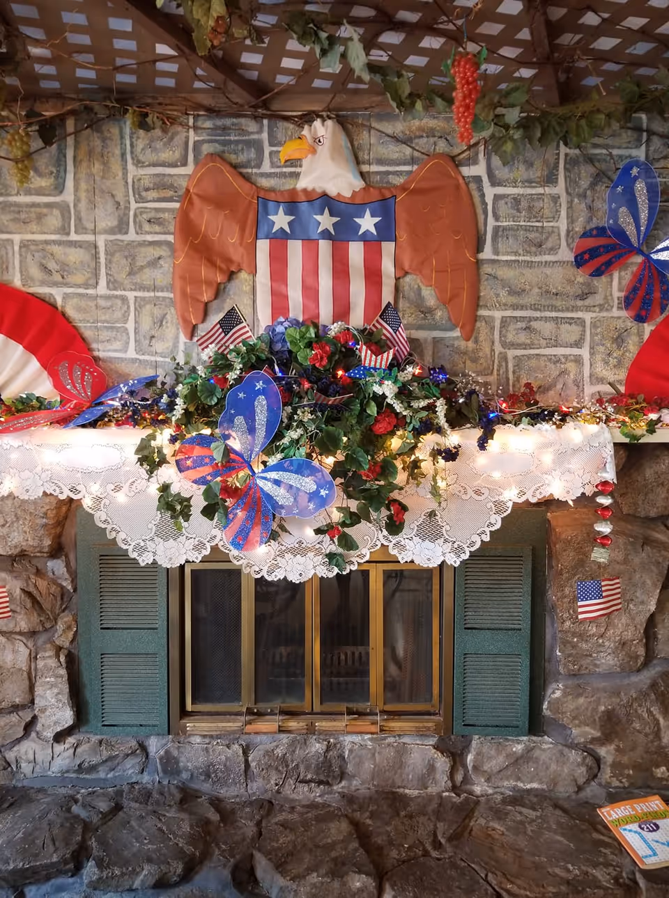 Stone fireplace mantel decorated with a patriotic eagle, American flags, red-white-blue butterflies, flowers and string lights.