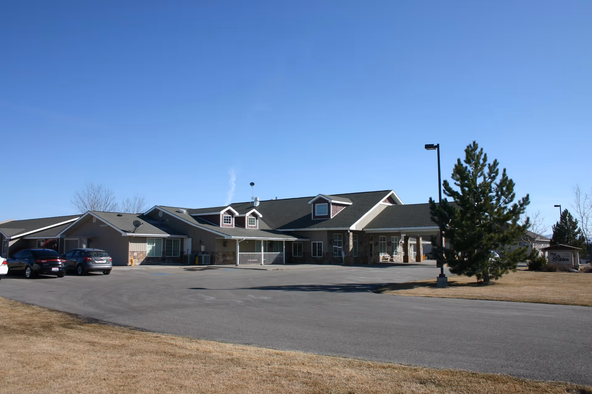 Single-story assisted living building with cars parked in front, a paved driveway and grassy lawn under a clear blue sky.