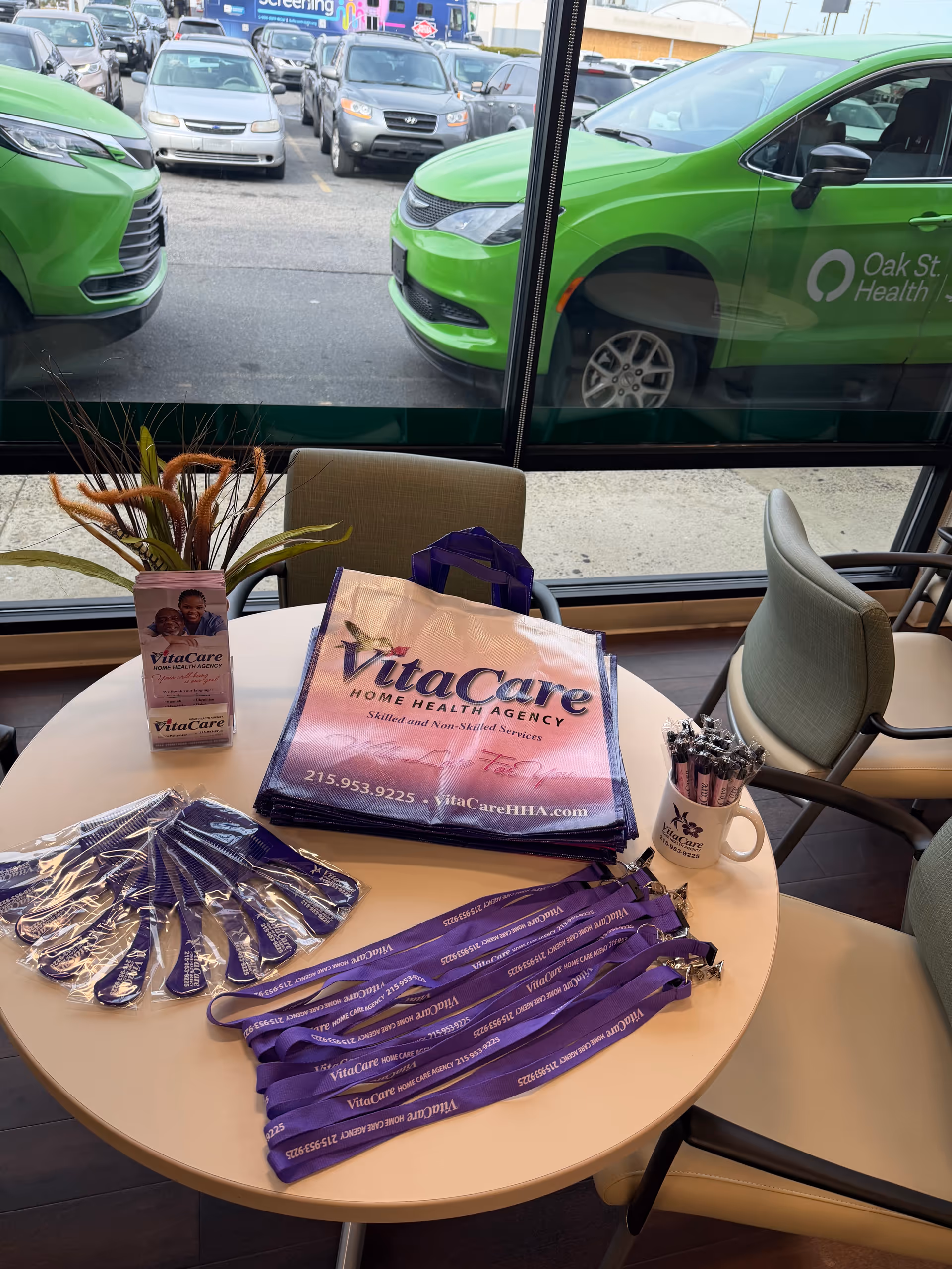 Indoor table setup with VitaCare promotional materials, purple lanyards, and pens by windows.