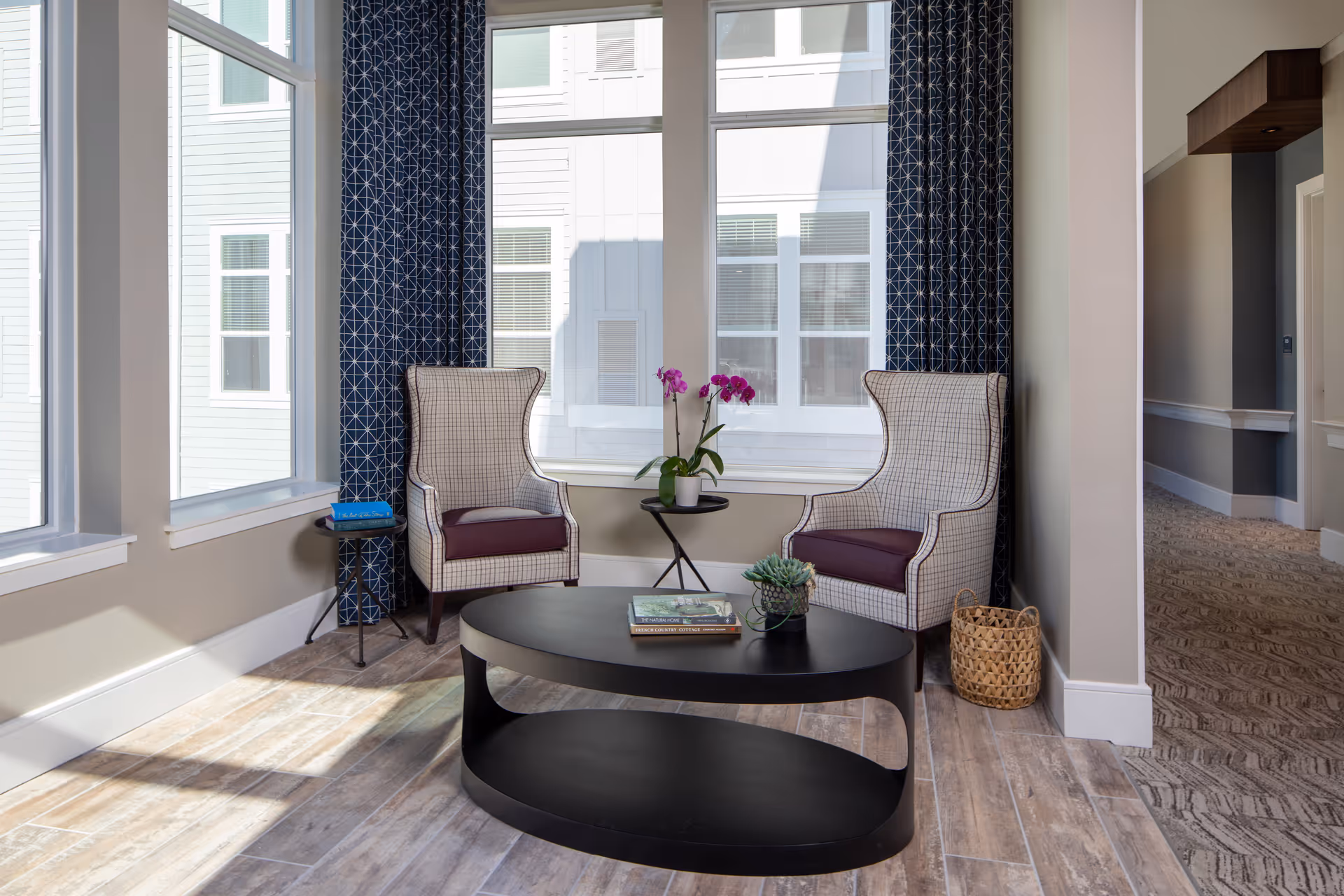 Sunlit lounge with two upholstered wingback chairs, a black oval coffee table with books and plants, and large windows with patterned curtains.