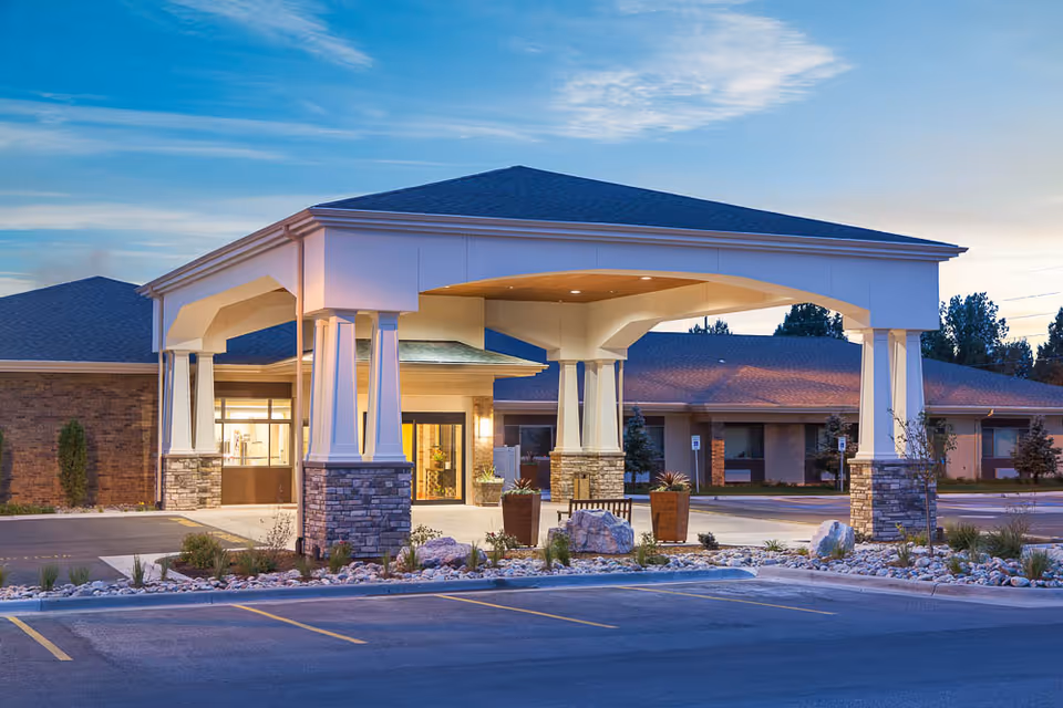 Covered entrance and porte-cochère of a senior living facility with stone pillars, landscaped rock beds, and a parking area at dusk.