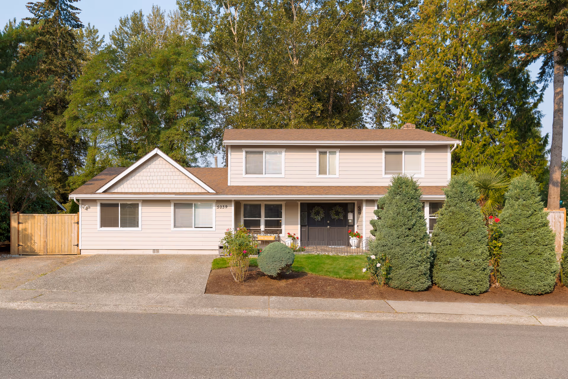 Two-story beige adult family home with a manicured front yard and tall trees.