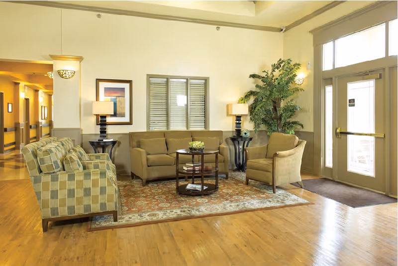 A well-lit sitting area in a nursing and rehabilitation facility featuring a patterned area rug, a beige sofa, two beige armchairs, and a checkered armchair. There are two side tables with lamps, a round coffee table with books and a small plant, a large potted plant in the corner, framed artwork on the wall, and a glass door entrance with windows allowing natural light.