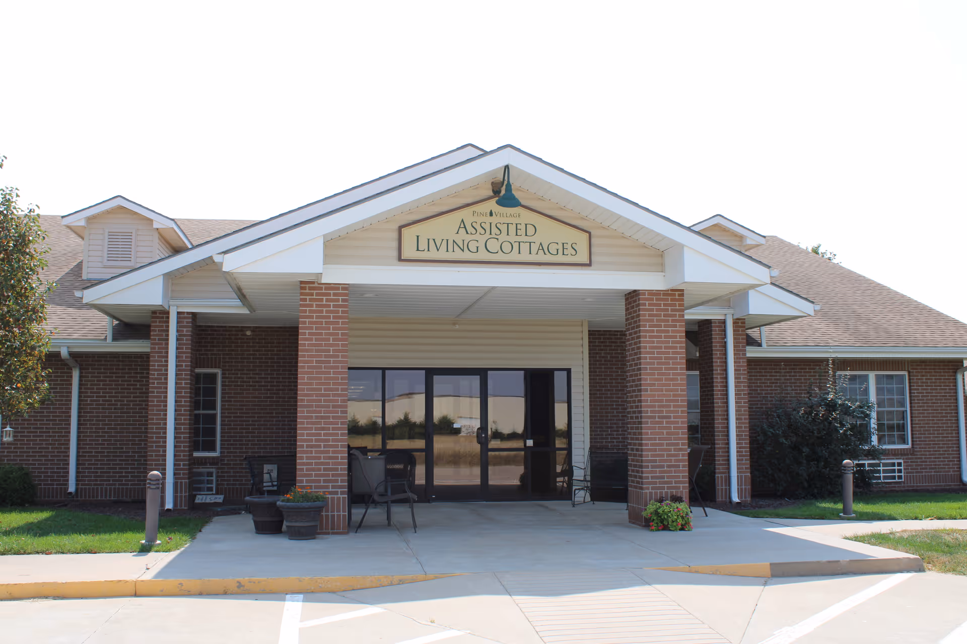 Front entrance of the Pine Village Assisted Living Cottages with a covered portico supported by brick columns and glass doors.