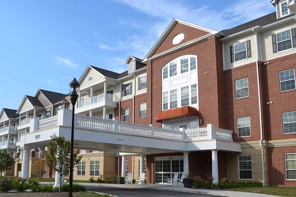 Exterior view of a multi-story senior living facility building with red brick and beige siding, featuring balconies, large windows, a covered entrance with white columns, and a street lamp in front under a clear blue sky.