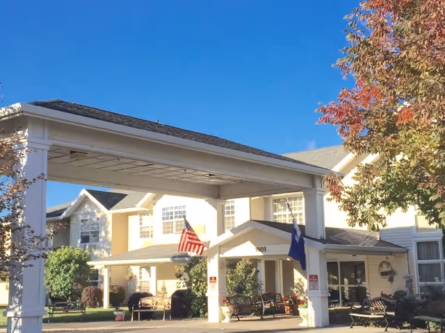 Entrance canopy and front facade of a light-yellow assisted living building with flags, outdoor benches, and autumn trees under a clear blue sky.