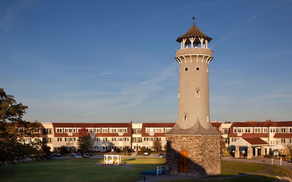 A tall stone and wood tower with a conical roof stands in front of a large three-story building with many windows and a red roof. The building is surrounded by a well-maintained lawn with a white gazebo and some trees under a clear blue sky.