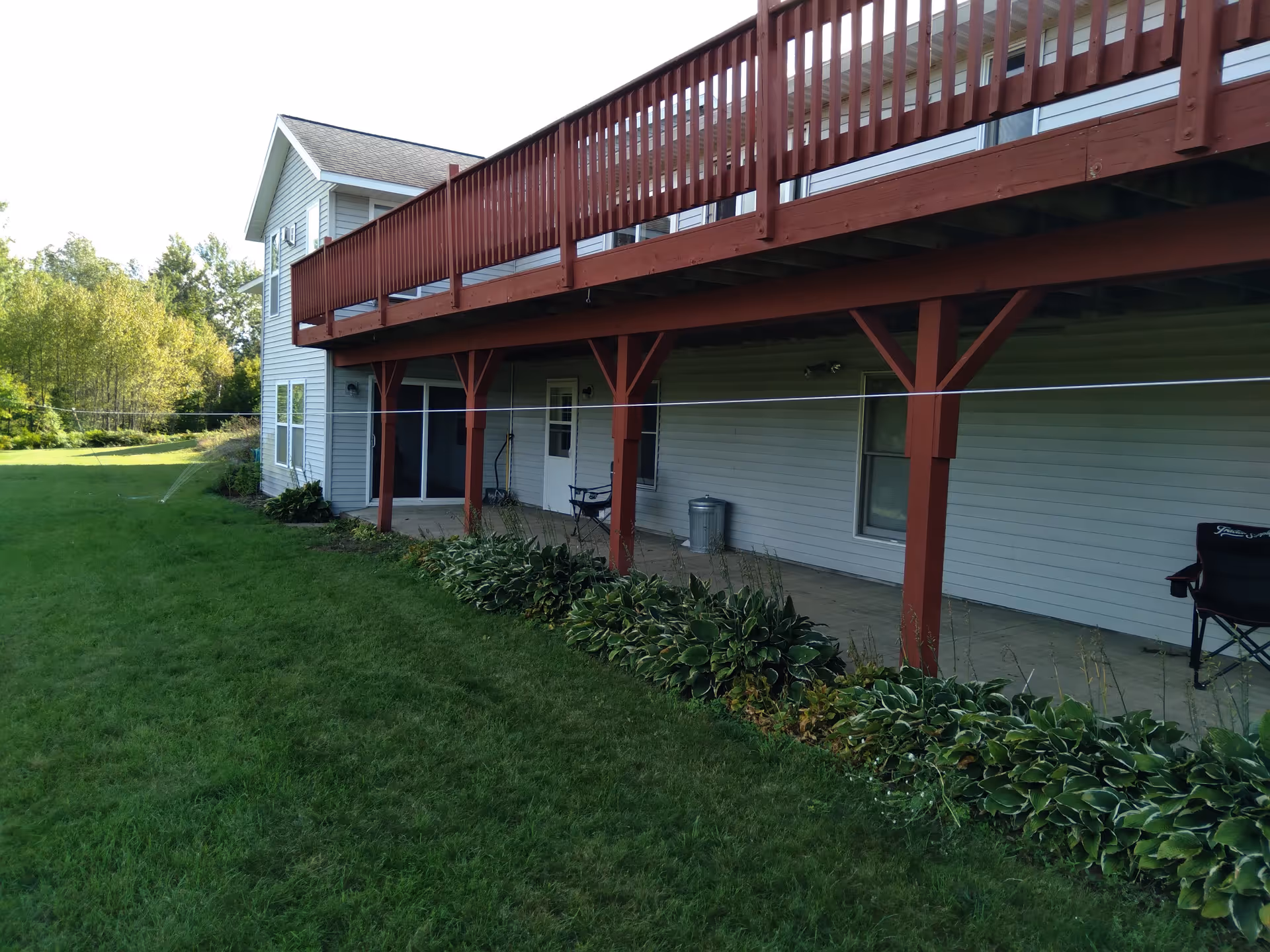 View of the back side of a two-story building with a large elevated wooden deck supported by red wooden beams. Below the deck is a concrete patio area with some chairs, a trash can, and plants along the edge. The building has white siding and several windows. There is a grassy lawn and trees in the background.