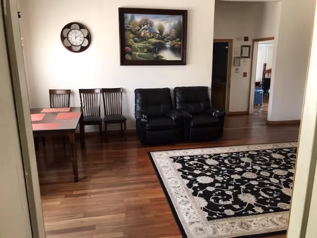 Interior view of a room with wooden flooring, a black and white patterned area rug, two black recliner chairs, three wooden chairs, and a wooden table with red placemats. A decorative wall clock and a framed landscape painting hang on the white wall.