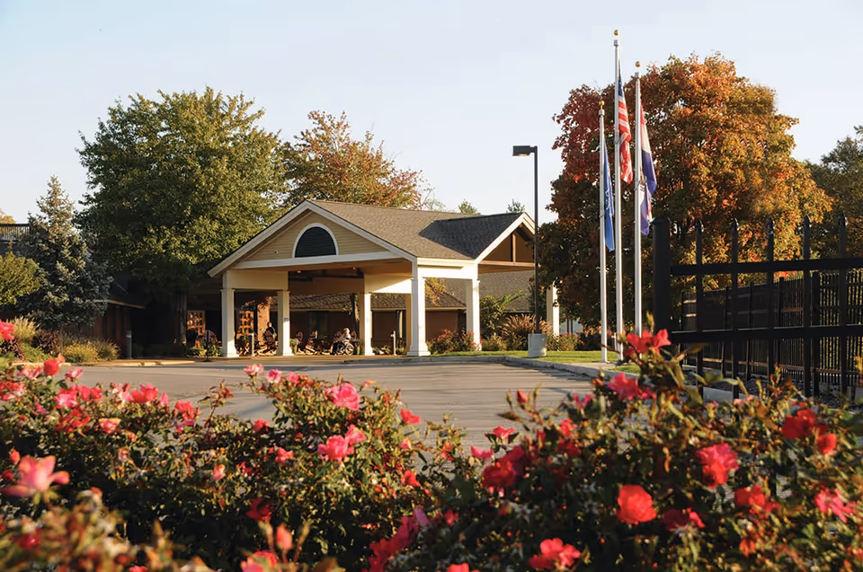 A facility entrance with a porte-cochere, flagpoles, and colorful flowerbeds in foreground.