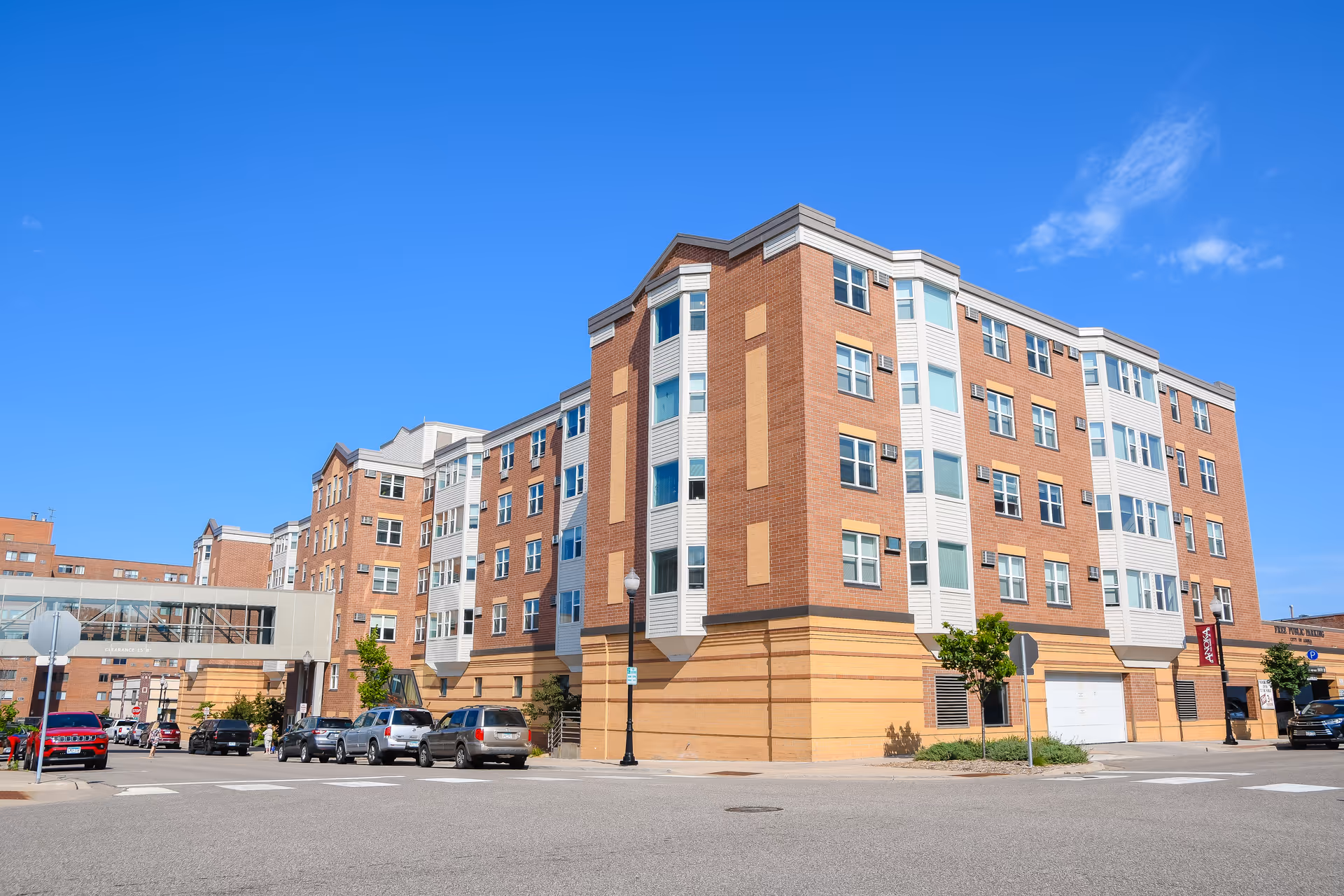 Multi-story brick and siding apartment building with a pedestrian skywalk, street parking, and a clear blue sky.