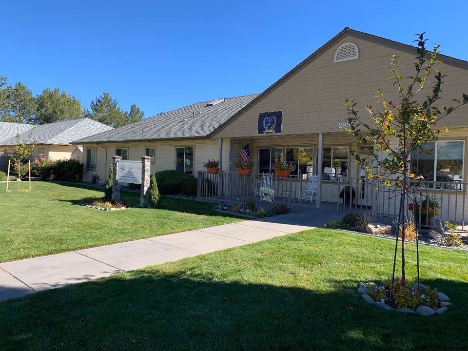 Single-story senior living facility with a covered front porch, railing, potted plants, and a green lawn.