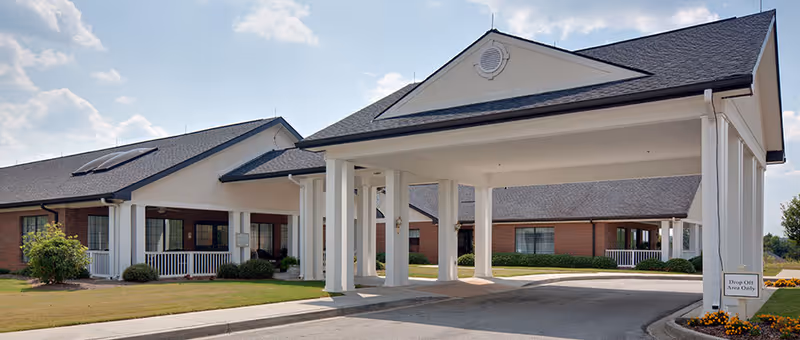 Exterior view of a single-story senior living facility with a covered drop-off area supported by white columns. The building has brick walls, white trim, and a dark shingled roof with solar panels. There is a small garden with flowers near the driveway and a sign indicating 'Drop Off Area Only'.