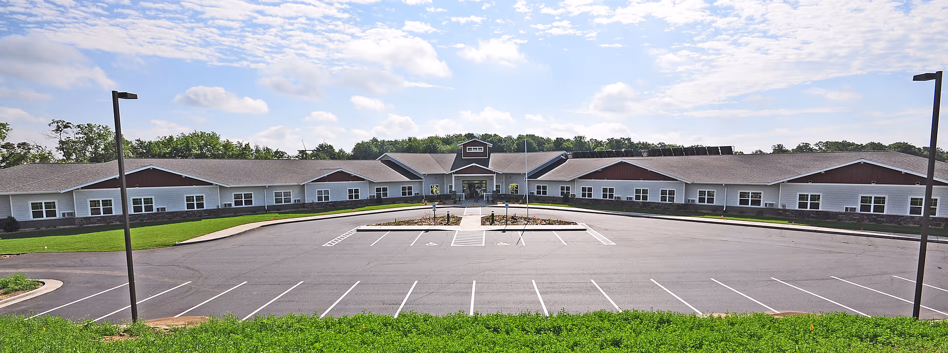 Wide exterior view of a single-story assisted living facility building with a large empty parking lot in front, surrounded by green grass and trees under a partly cloudy sky.