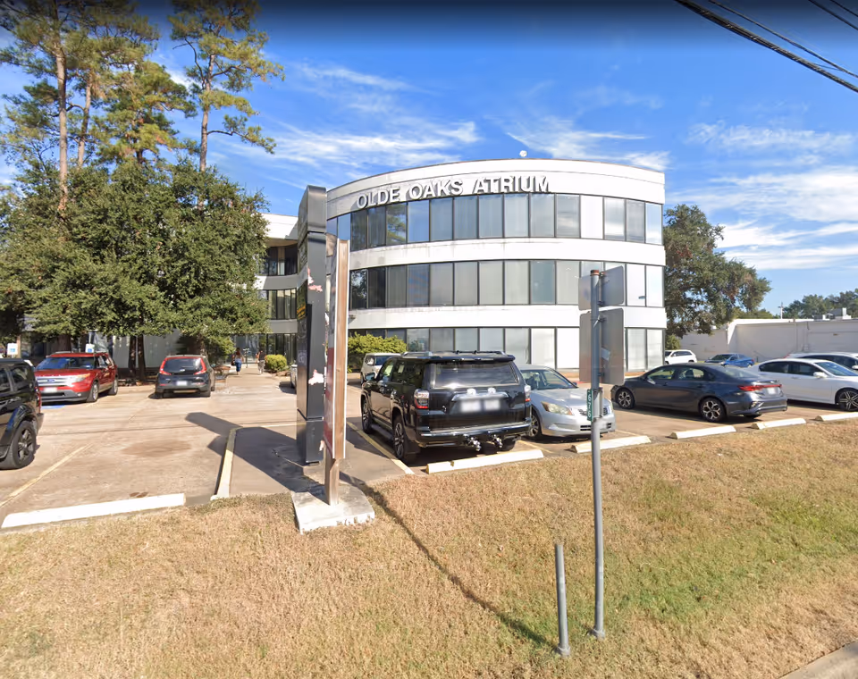 Curved glass front of Olde Oaks Atrium building with parked cars and trees nearby.