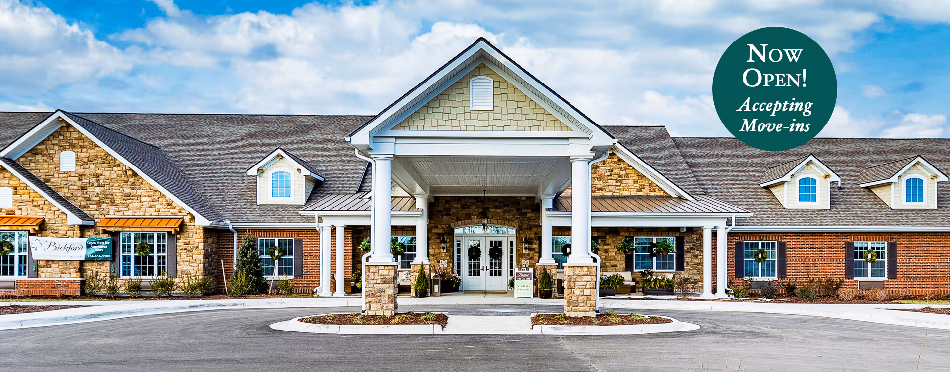 Front exterior view of Bickford of Canton facility with a large covered entrance supported by white columns, brick and stone facade, multiple windows with wreaths, and a circular driveway. A green circular sign on the right side reads 'Now Open! Accepting Move-ins'.