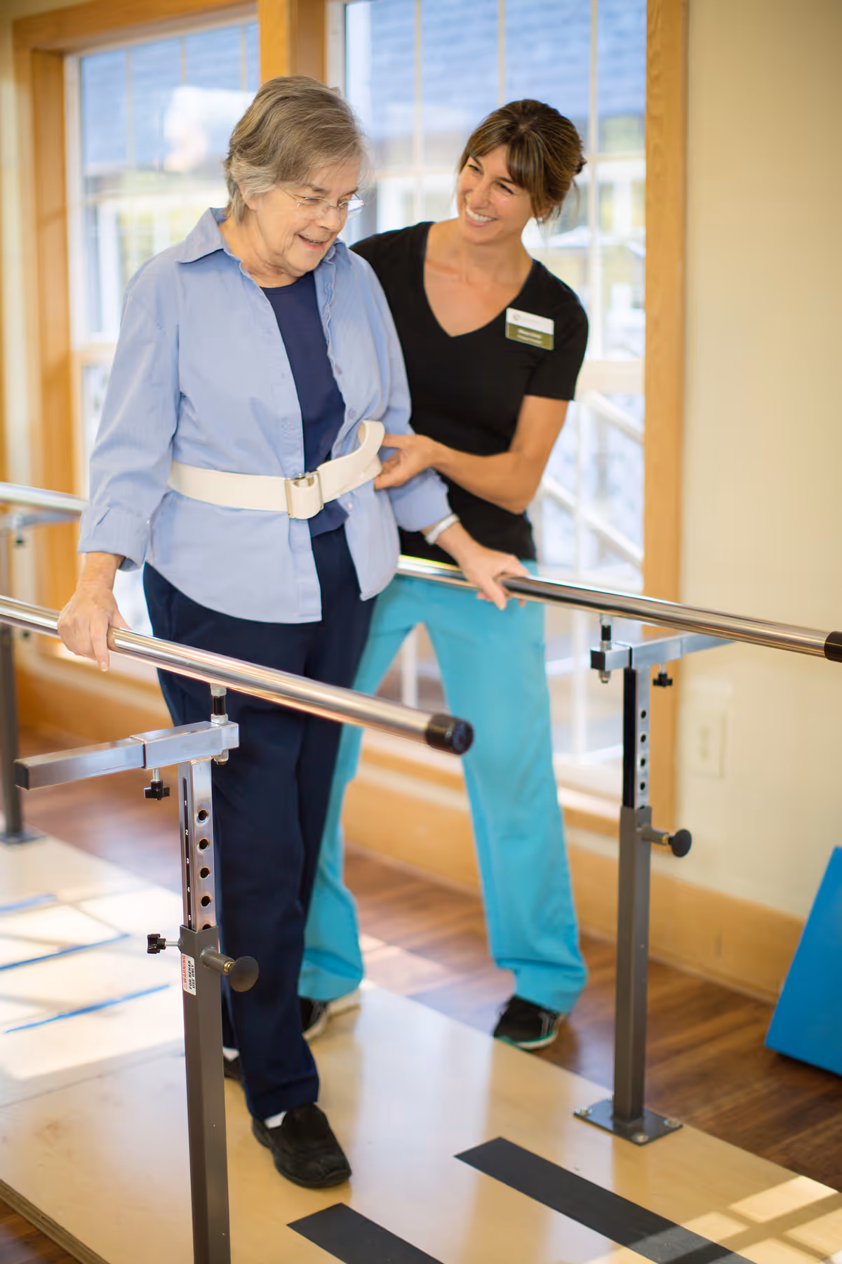 An elderly woman wearing a safety belt is walking between parallel bars for physical therapy, assisted by a smiling female healthcare worker in a black shirt and turquoise pants inside a well-lit room with wooden floors and large windows.