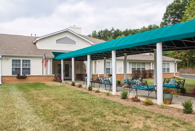 Exterior view of a single-story senior living facility building with a covered outdoor seating area featuring metal benches with teal cushions. The building has white siding with brick accents, multiple windows, and an American flag near the entrance. The area is surrounded by grass and some small plants.