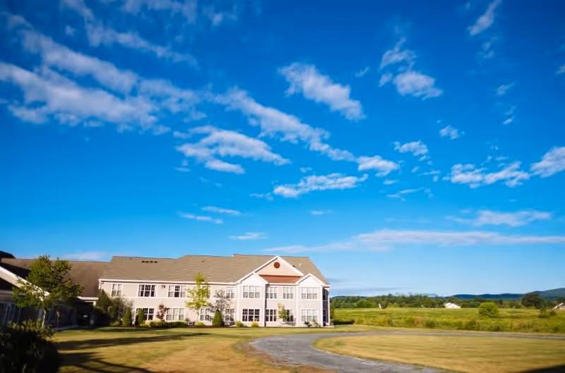 A large two-story building with multiple windows set in a spacious green field under a bright blue sky with scattered clouds. The building is surrounded by a well-maintained lawn and a curved gravel driveway.