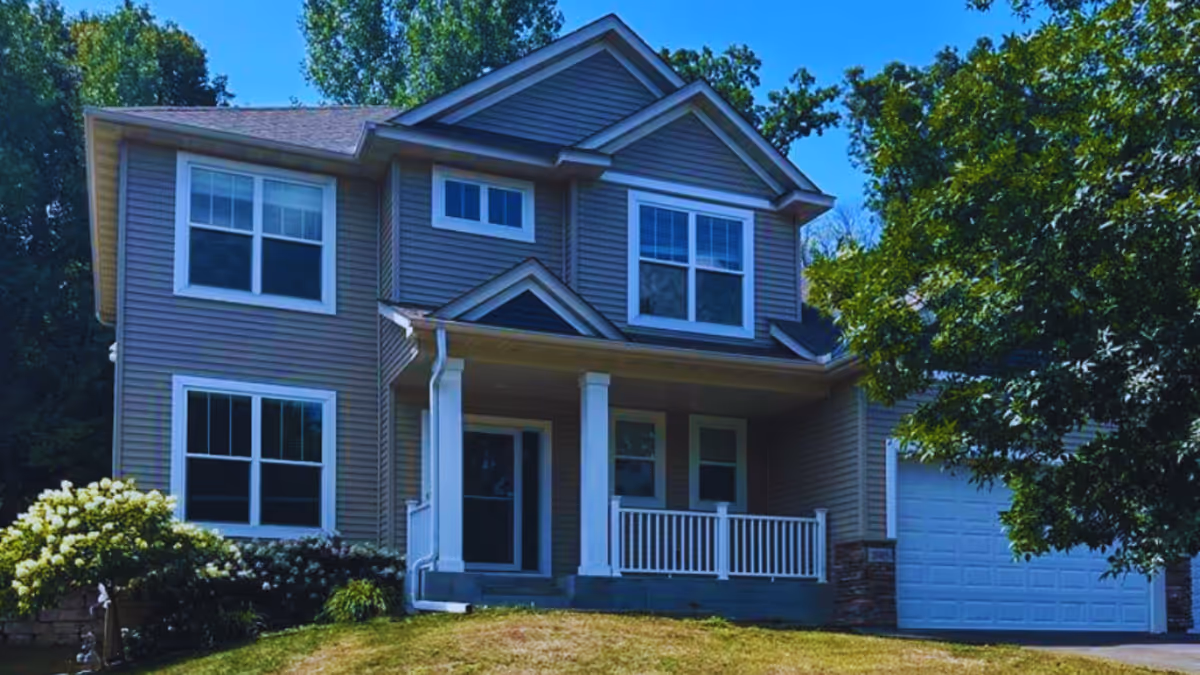 Two-story beige house with front porch, white trim, and surrounding trees.