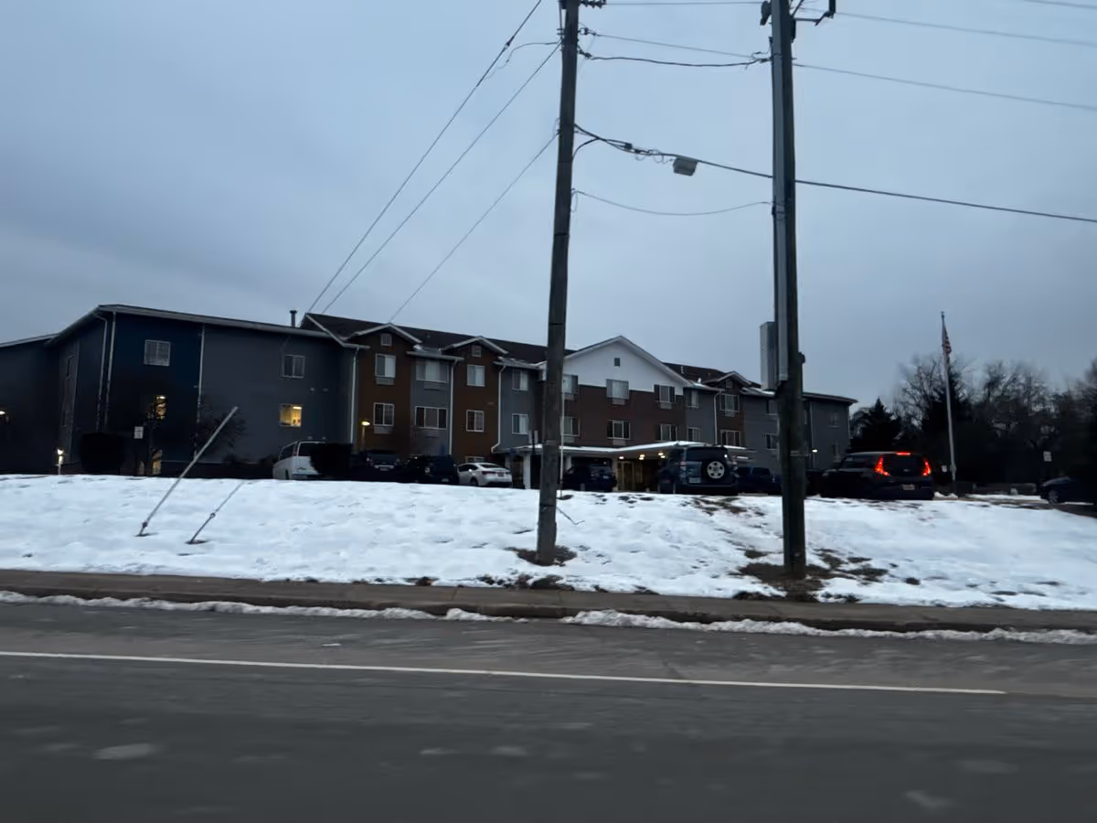 Three-story senior living building with a snow-covered front lawn, parked cars, and utility poles in the foreground under an overcast sky.