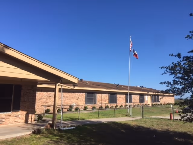 Single-story brick senior living facility exterior with flagpole, blue sky, and green lawn.