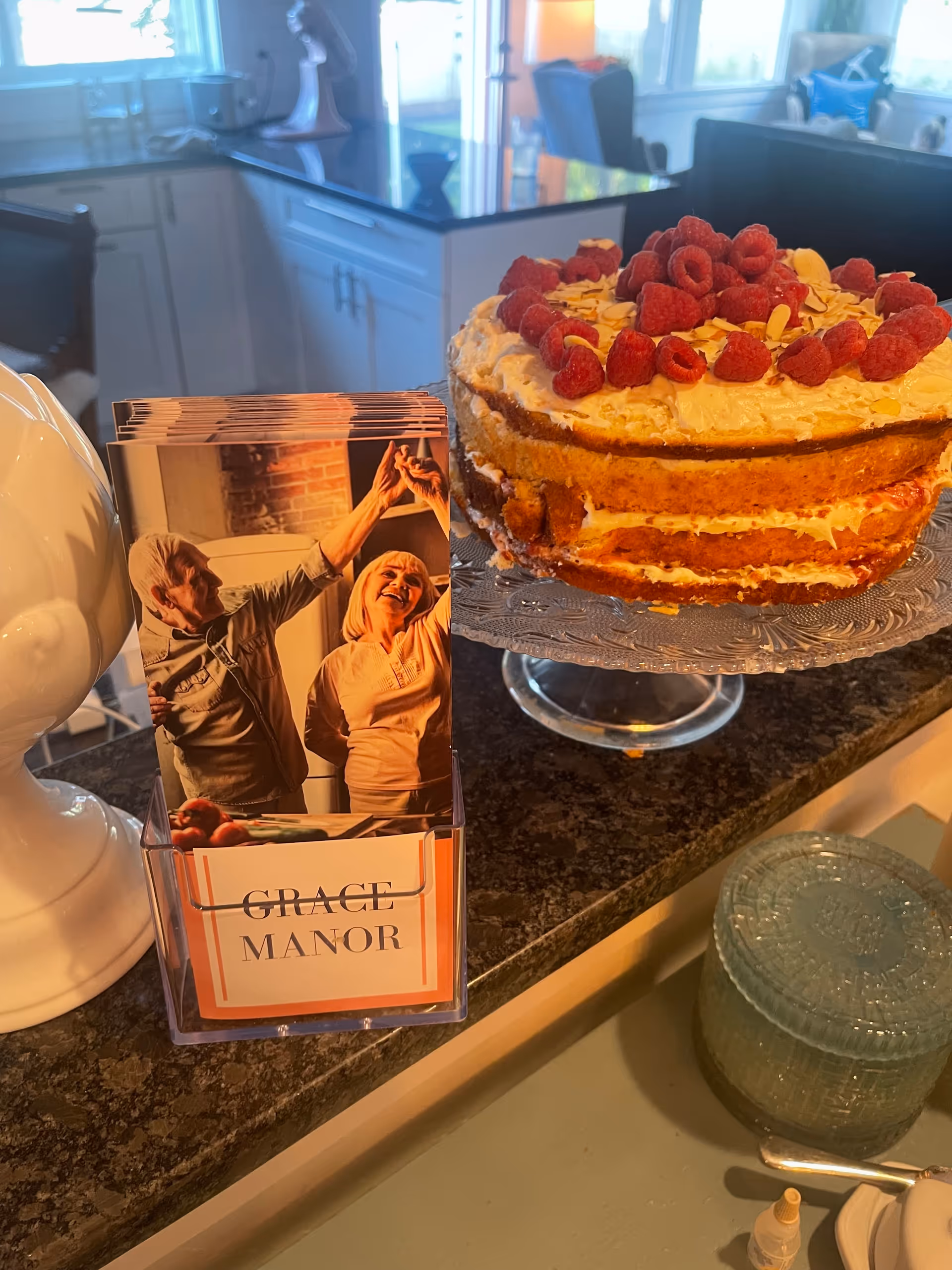 Cake with raspberries on a cake stand beside a Grace Manor brochure on a kitchen counter.