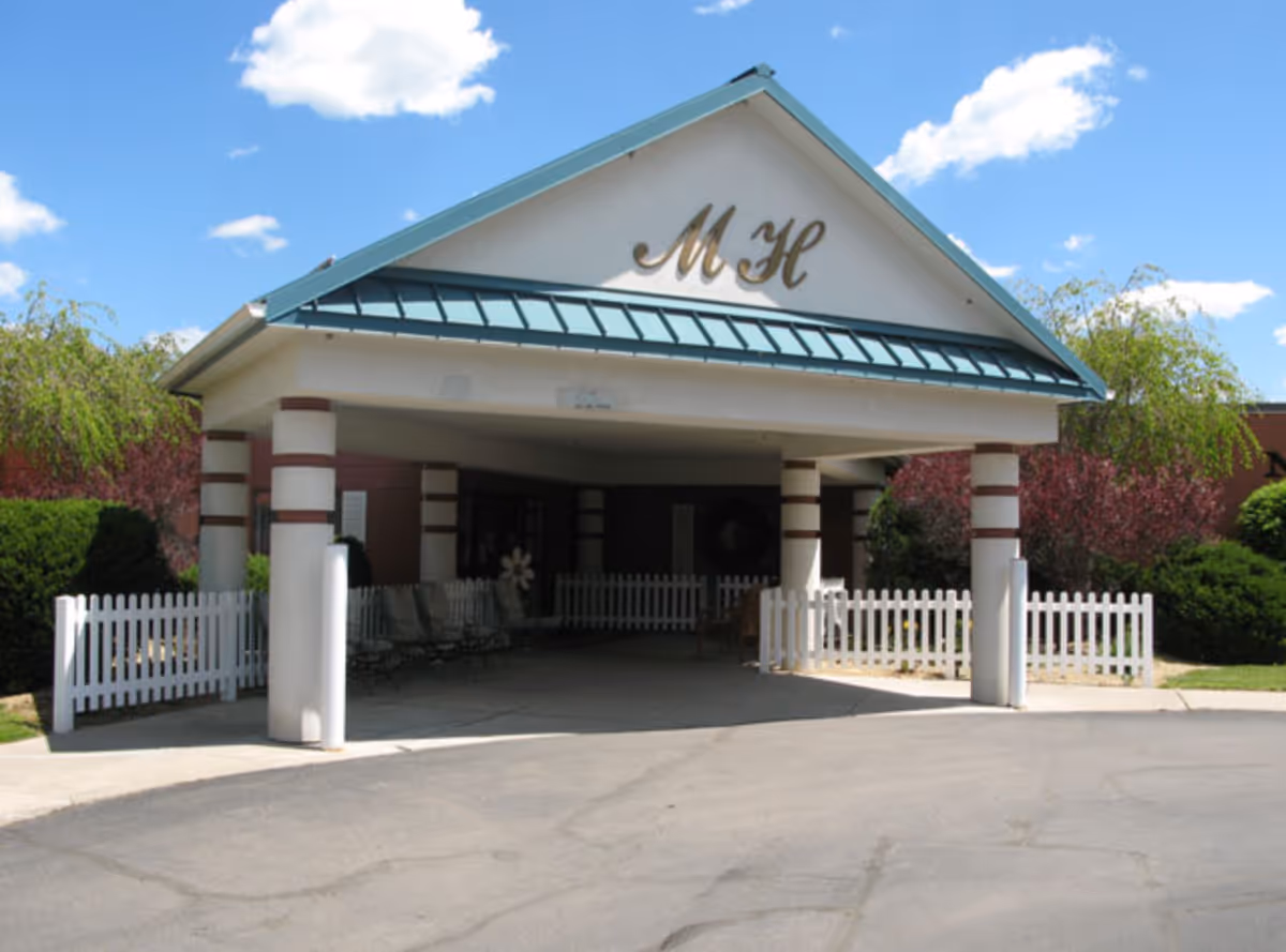 Covered front entrance of a senior living building with a teal metal roof, columns, white picket fencing, and 'MH' letters on the gable.