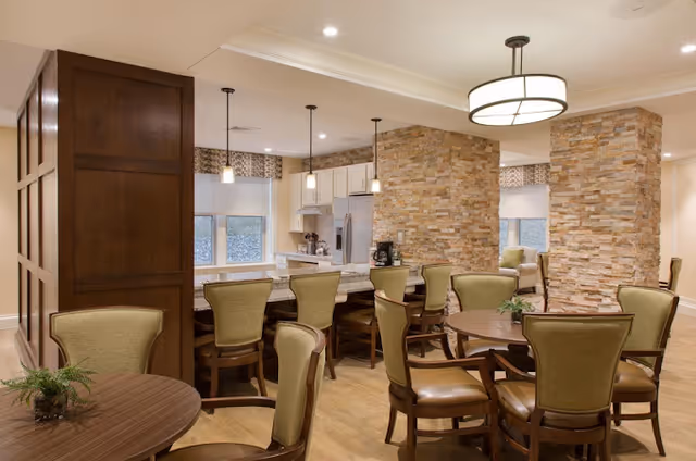 Communal dining area with round tables and upholstered chairs, a countertop with bar stools, and a kitchen behind stone pillars.