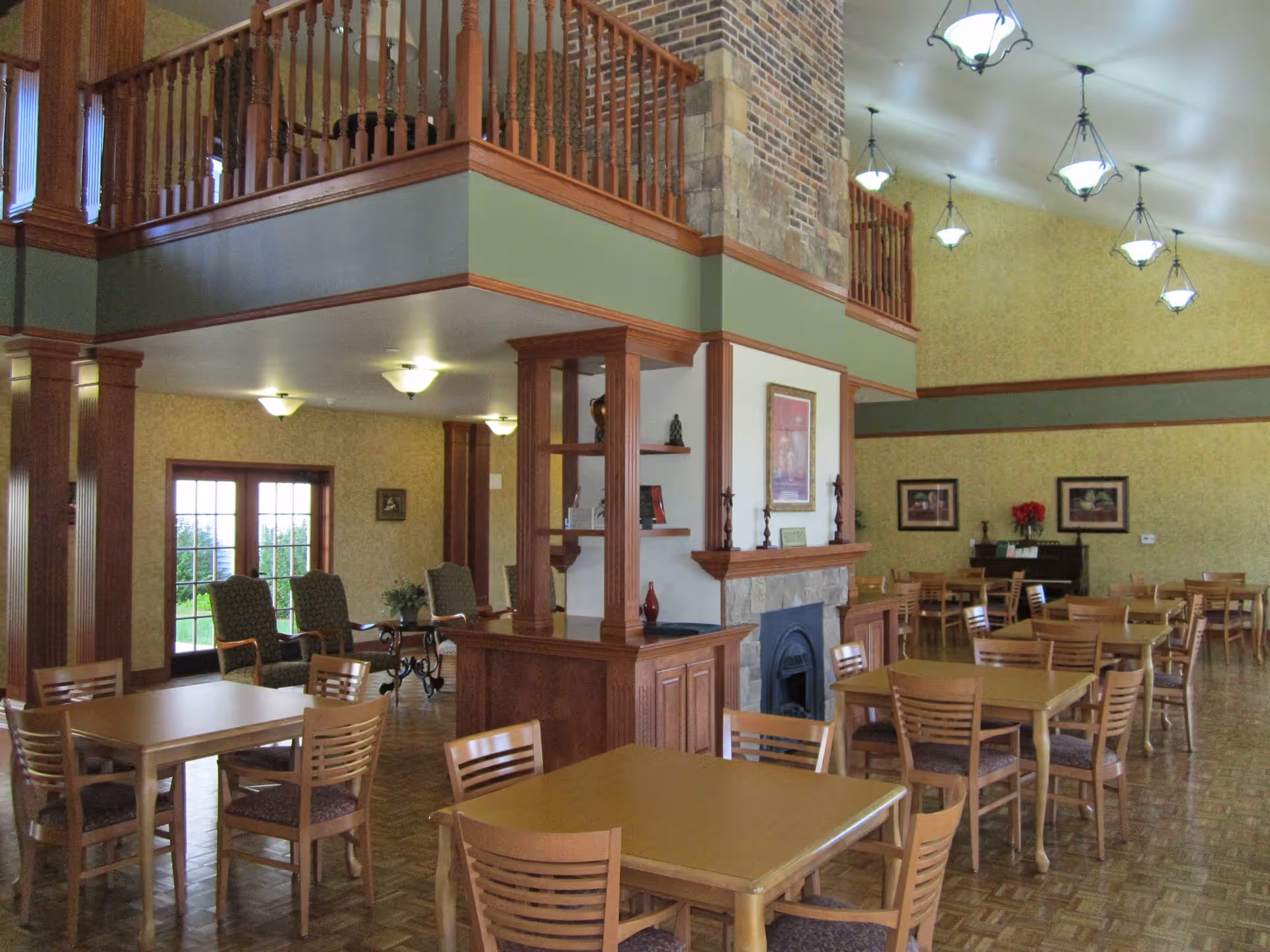 Spacious communal dining area with multiple wooden tables and chairs, a stone fireplace, high ceiling with hanging lights and an upper balcony.
