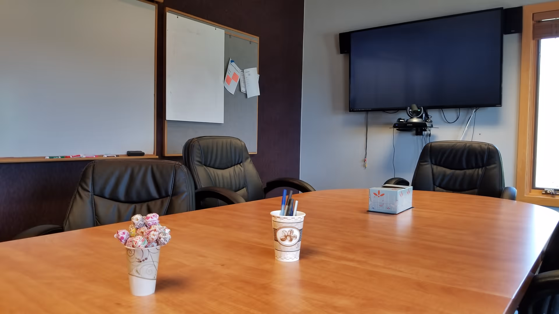 A conference room with a large wooden table, black chairs, a mounted TV, and whiteboards.