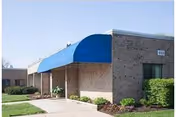Brick facility exterior with a blue curved awning over the entrance and landscaped path.