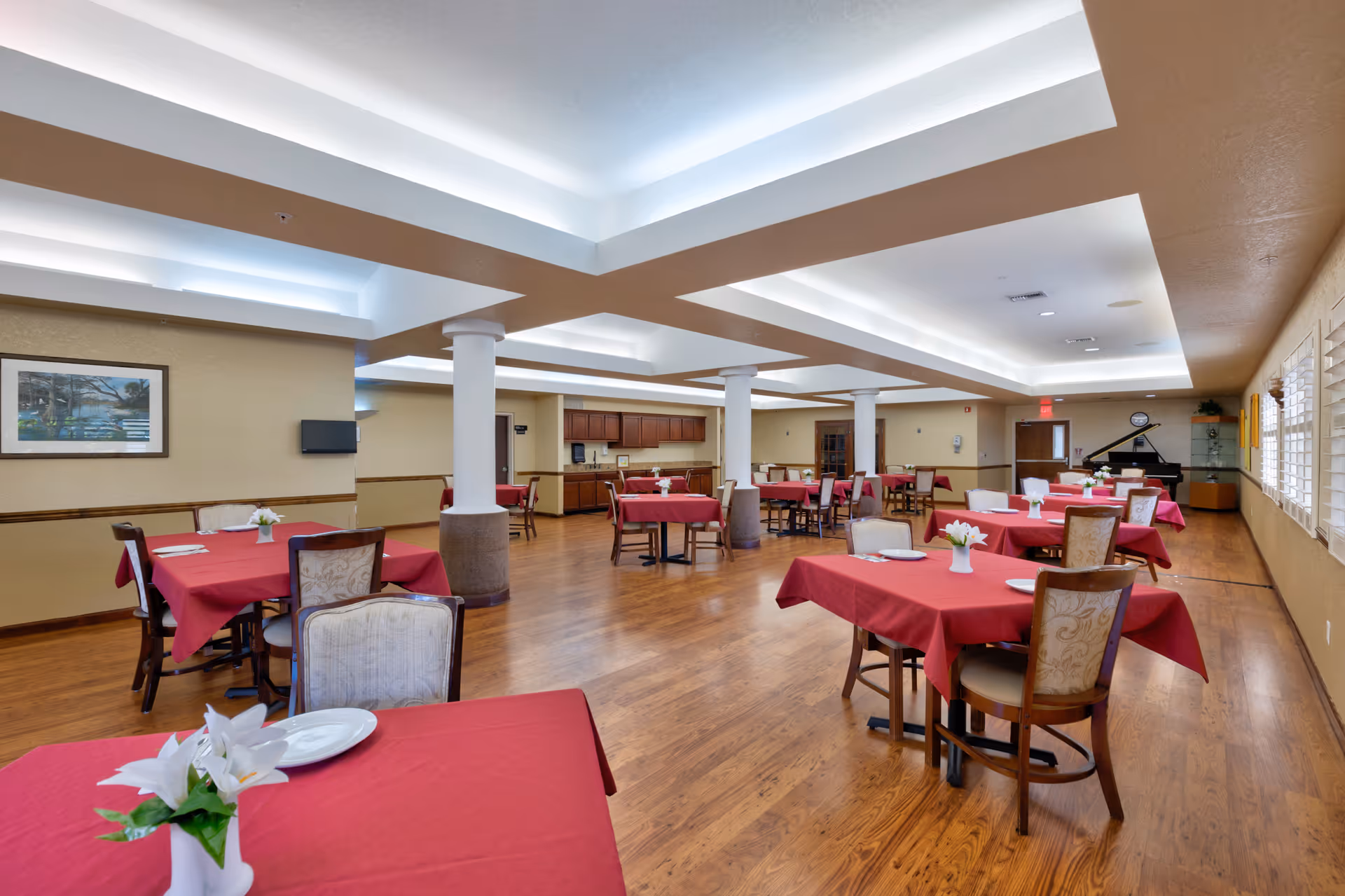 Spacious dining room with multiple tables covered in red tablecloths, each set with white plates and small white flower centerpieces. The room has wooden flooring, beige walls, white ceiling with recessed lighting, and several white columns. A piano is visible in the back corner near a door and windows with white blinds line one side of the room.
