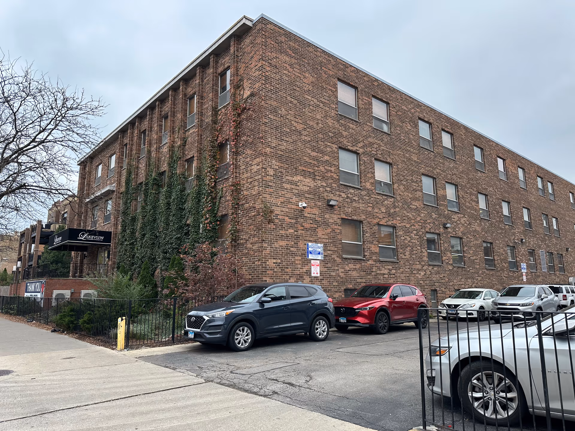 Three-story brick nursing facility with ivy-covered facade, an awning entrance and several cars parked in front.