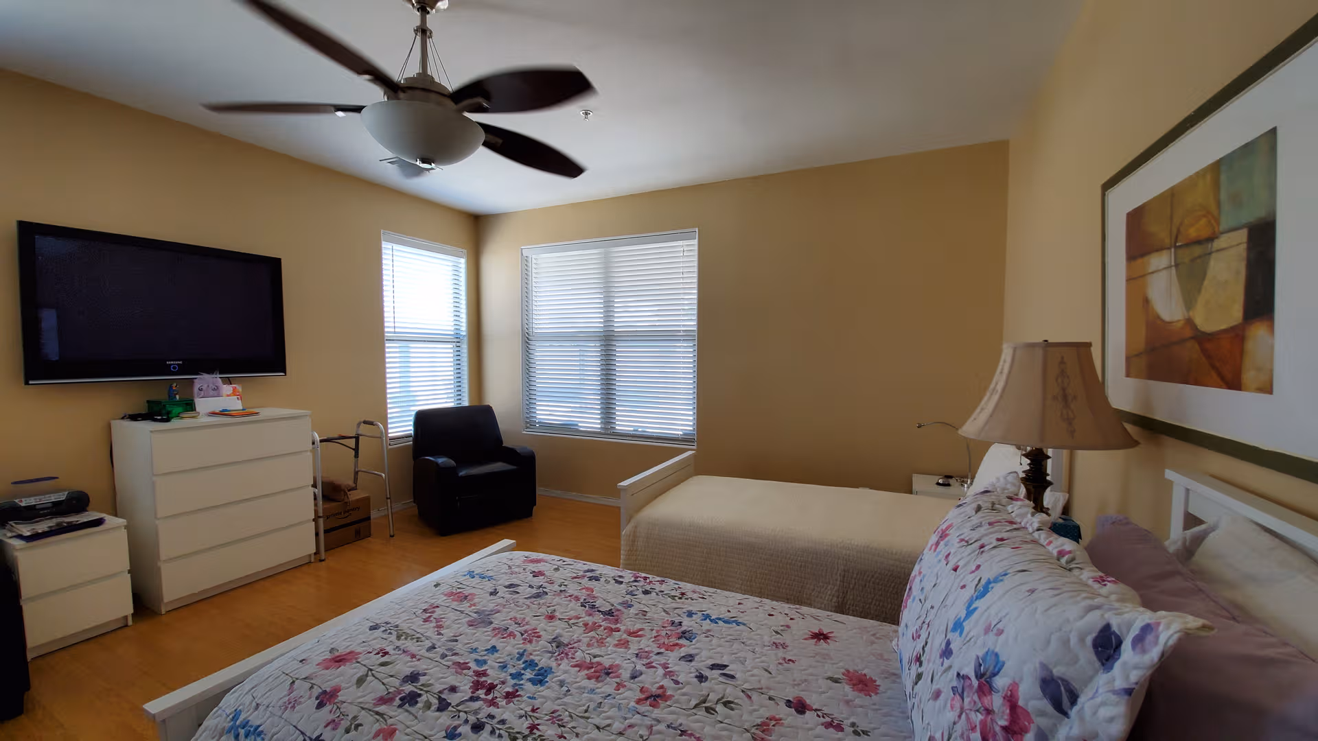 A spacious senior bedroom with two beds, floral quilts, dresser, TV, and ceiling fan.
