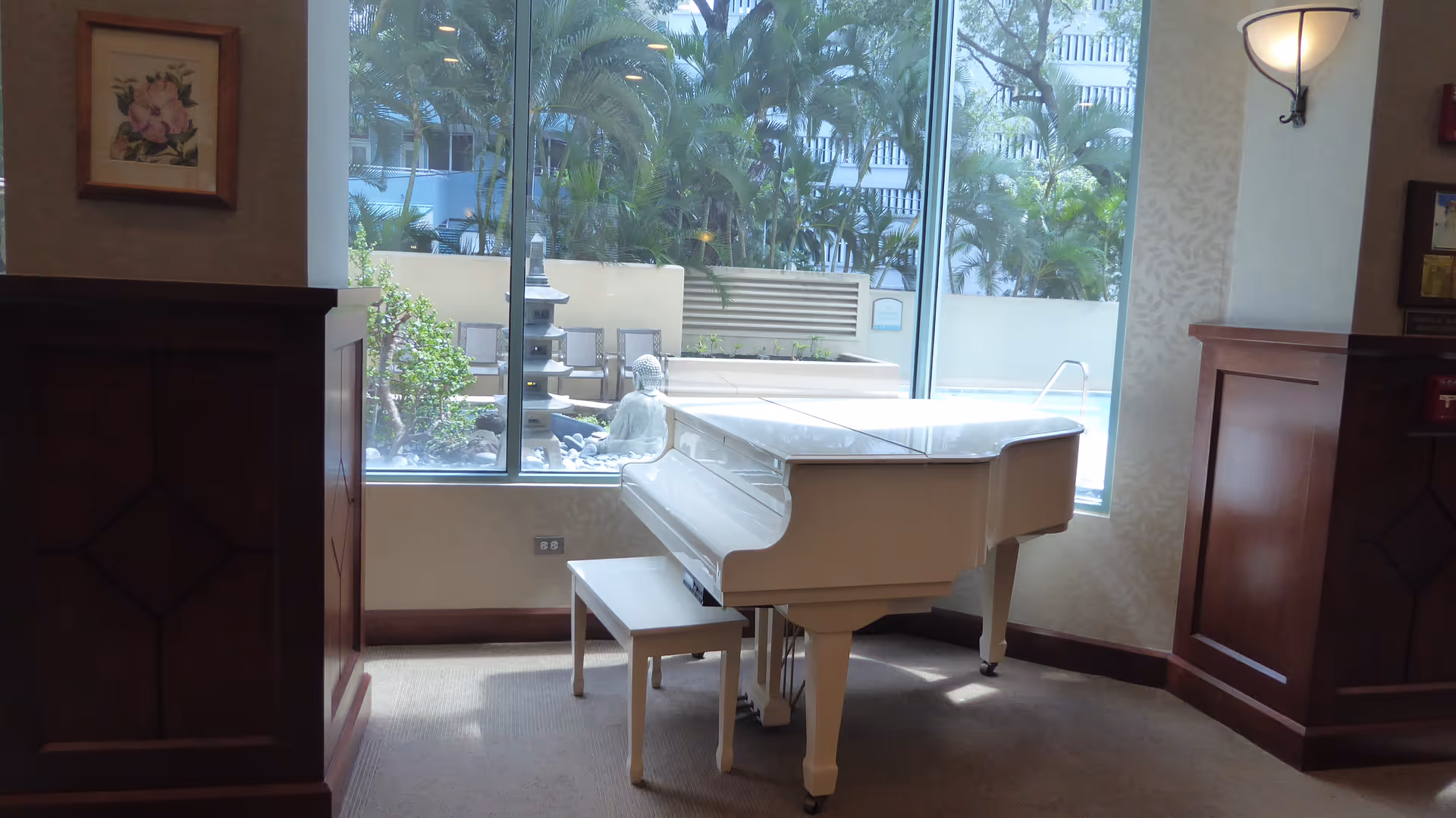 White grand piano with bench sits in a bright lobby-like room overlooking a pool