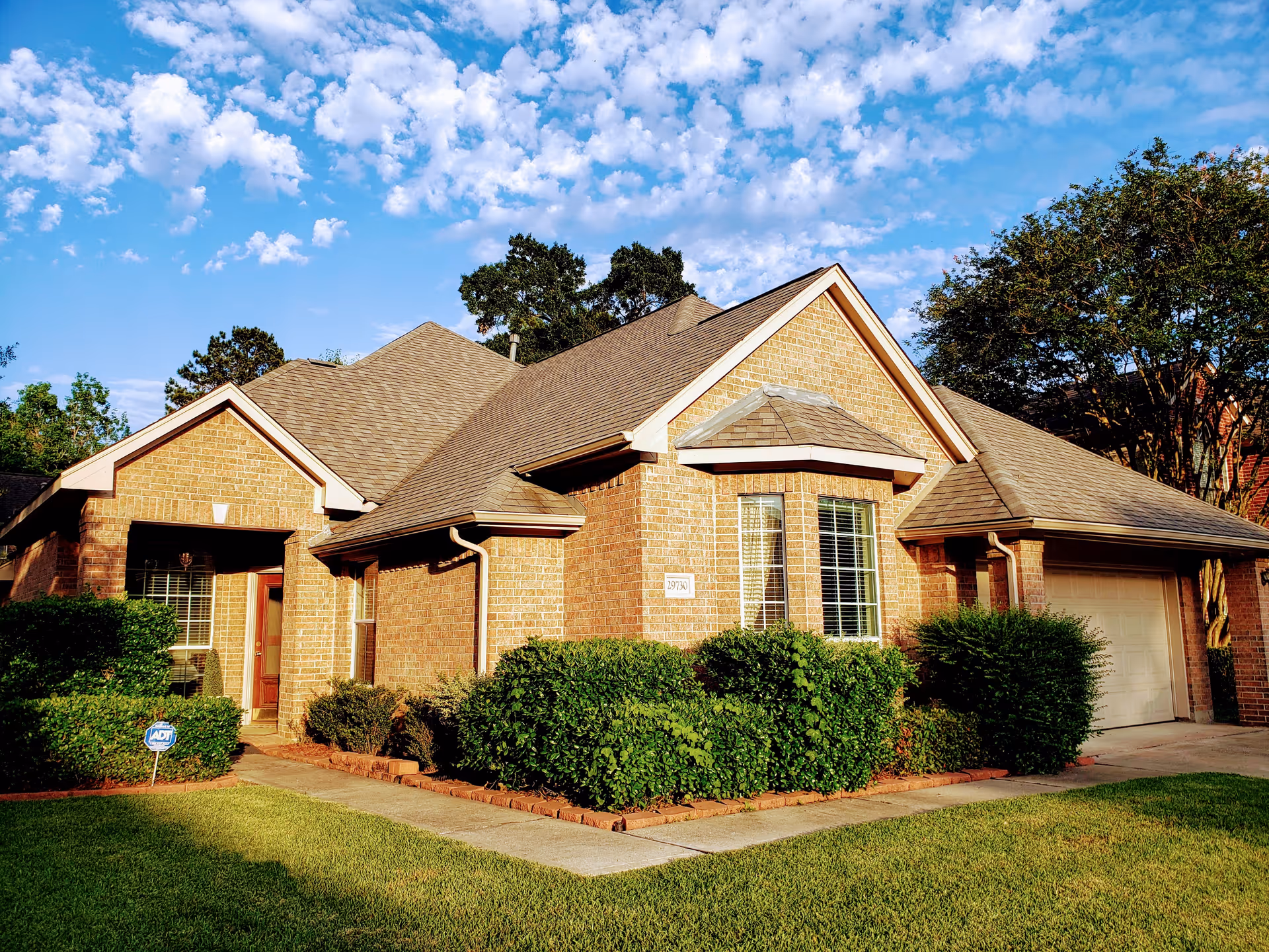Front exterior of a single-story brick house with a manicured lawn, shrubs, and an attached garage under a blue sky.