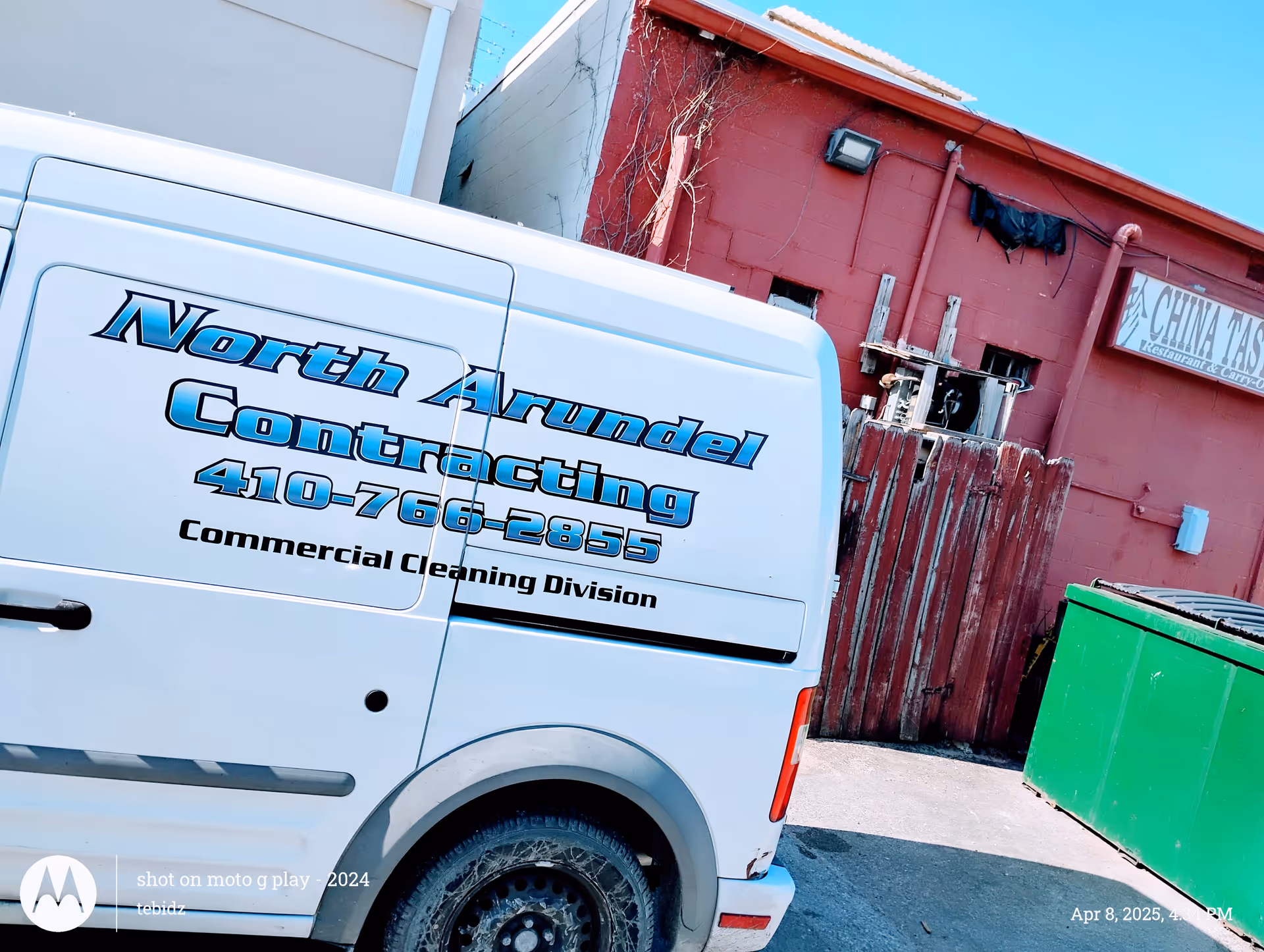 White van with North Arundel Contracting graphics parked beside a red building and green dumpster.