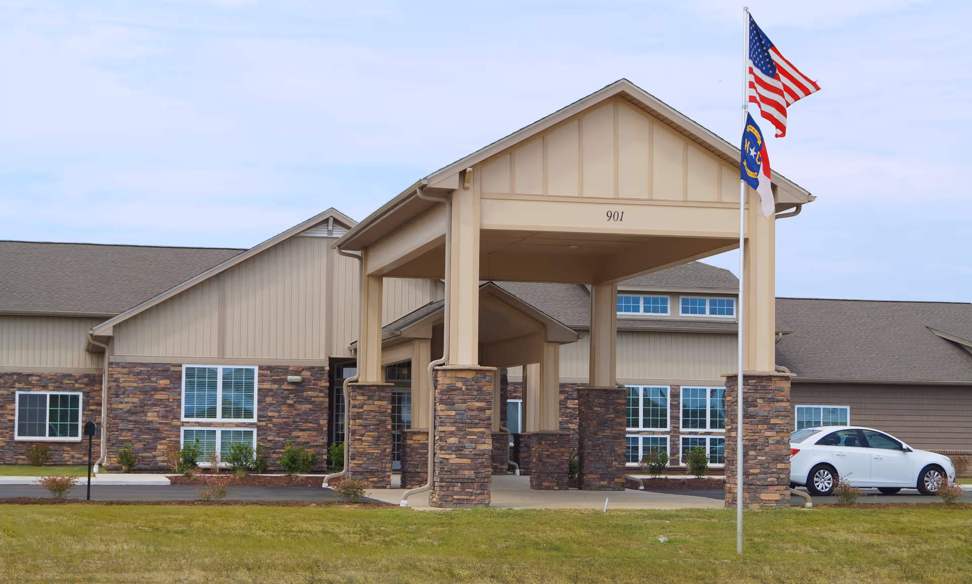 Exterior view of a senior living facility building with a covered entrance supported by stone pillars. Two flags, the American flag and a state flag, are flying on a flagpole in front of the building. A white car is parked near the entrance.