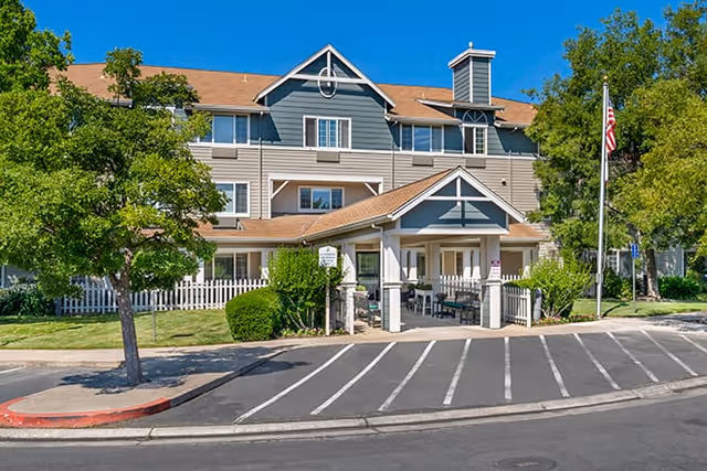 Exterior view of a multi-story senior living facility with a covered entrance, surrounded by trees and greenery. There is a white picket fence, a flagpole with an American flag, and a parking area in front.