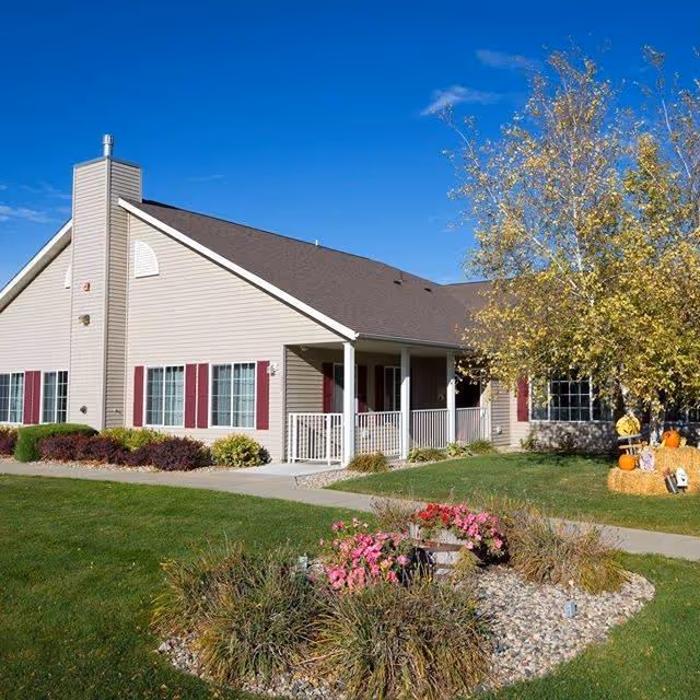Single-story beige senior living cottage with a front porch, landscaped lawn, and seasonal pumpkin decorations under a blue sky.