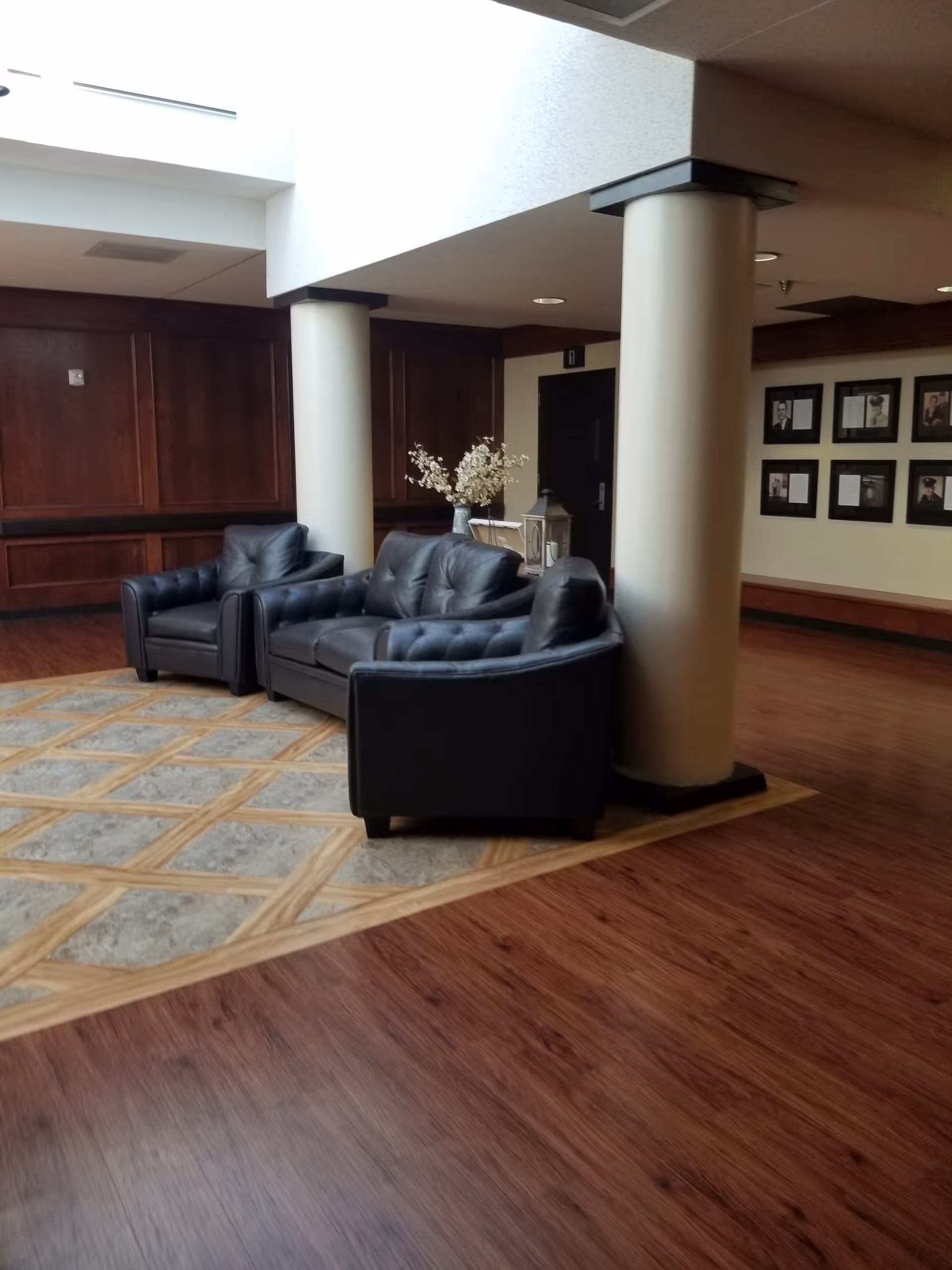 Lobby seating area with black leather sofas, pillars, and wood floors in a senior facility.