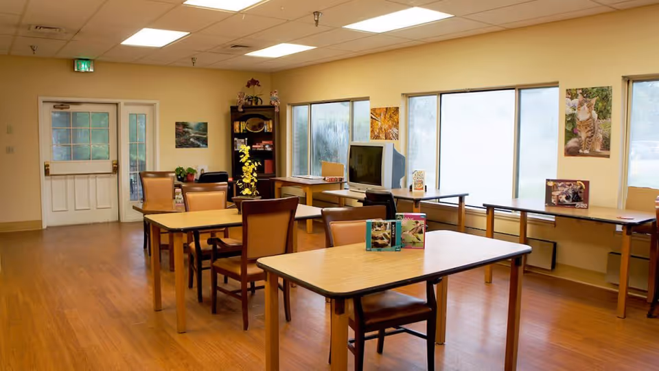 A well-lit room with wooden floors and beige walls featuring several tables and chairs arranged in rows. There is a TV on a table near the windows, and a bookshelf with decorative items and books. The windows have frosted glass, and there are pictures of cats and nature on the walls. A white door with glass panels is visible on the left side of the room.
