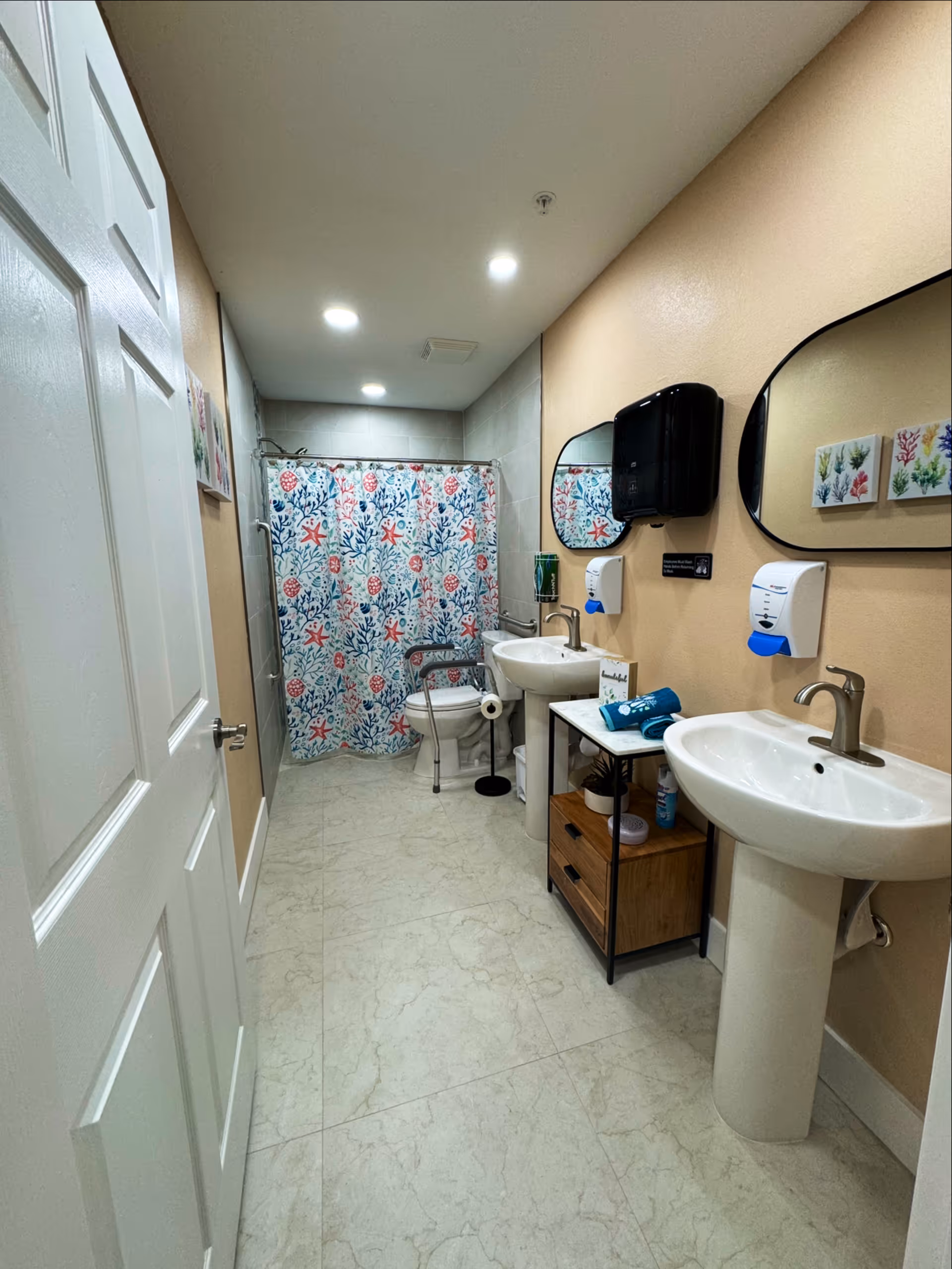 Long bathroom with dual sinks, mirrors, soap dispensers, and a tropical-pattern shower curtain.