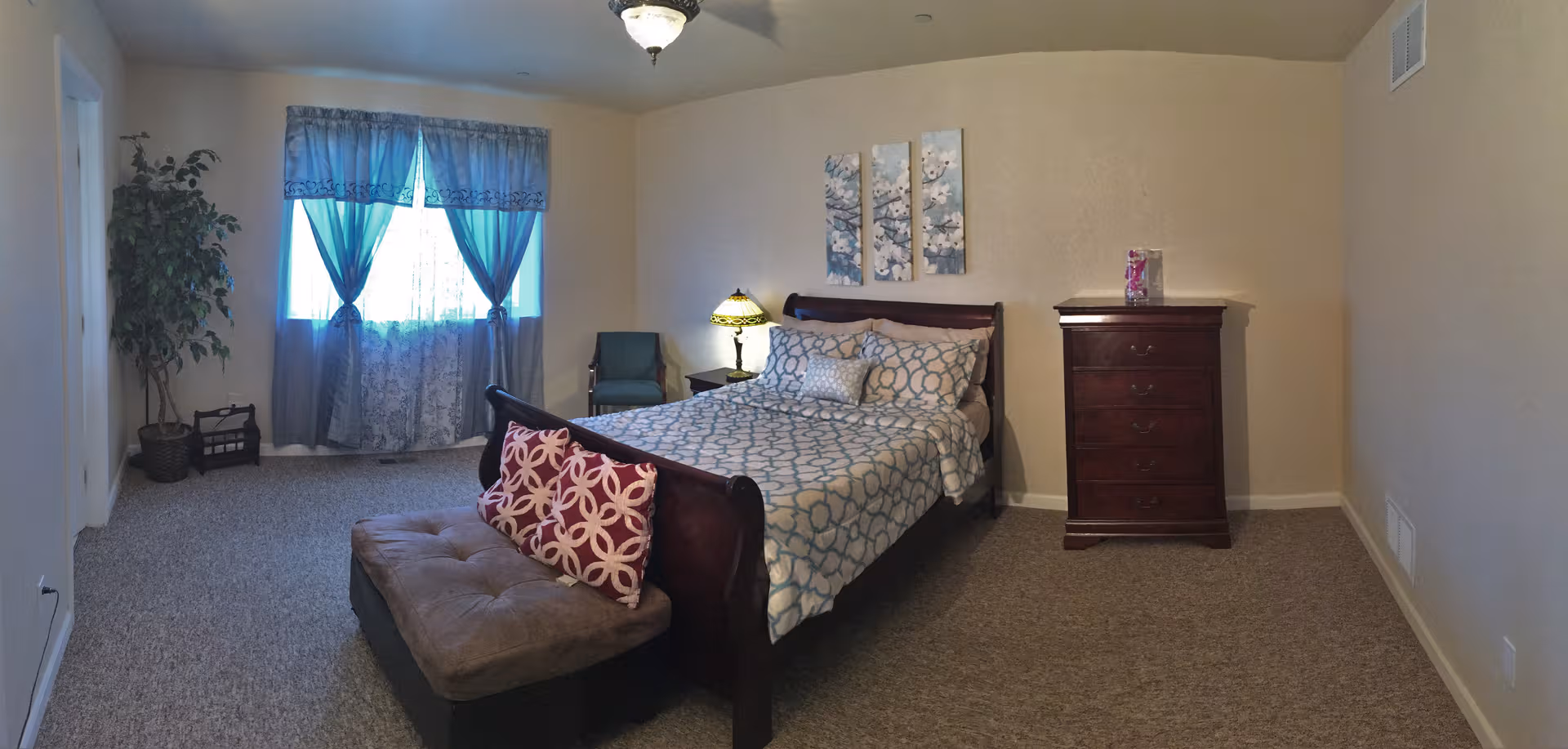 A bedroom in Pine Ridge Terrace Assisted Living with a bed, dresser, plants, and window.