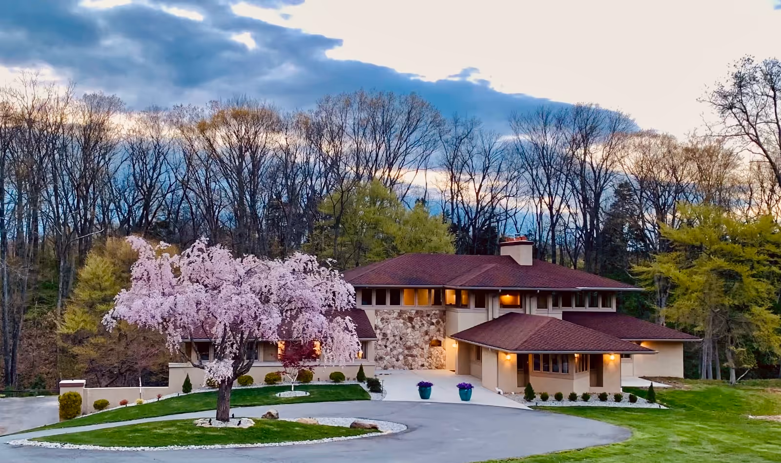 A large, modern house with a reddish-brown roof surrounded by a well-maintained lawn and landscaping. In front of the house, there is a blossoming tree with pink flowers. The house is set against a backdrop of tall trees with bare branches under a cloudy sky at dusk.